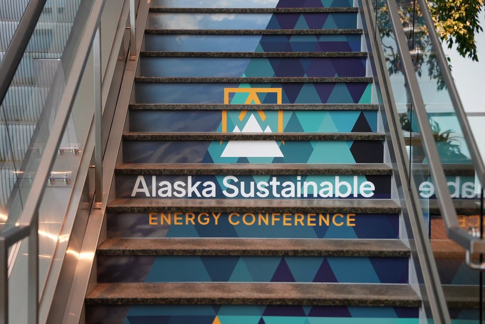A vibrant staircase features a mural on its risers with geometric patterns in shades of blue and green, a white mountain silhouette, and the words 'Alaska Sustainable Energy Conference' in white and orange lettering.