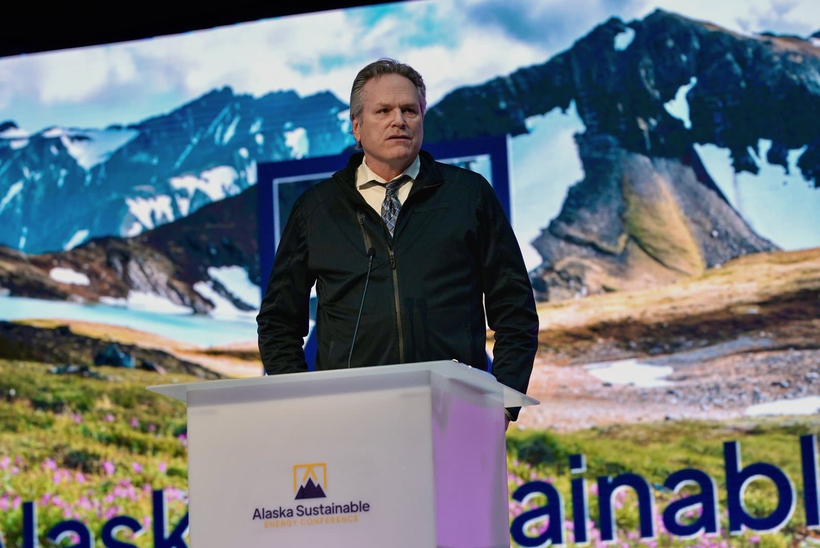 A man in a black jacket and tie stands at a podium with the 'Alaska Sustainable Energy Conference' logo. He is speaking in front of a large screen displaying a scenic Alaskan mountain landscape with snow, water, and green fields.
