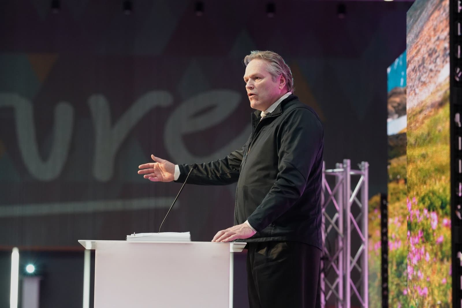 A man in a black jacket and white shirt speaks at a podium, gesturing with his left hand, during the Anchorage Sustainable Energy Conference. Large screens behind him display event graphics and a vibrant landscape.