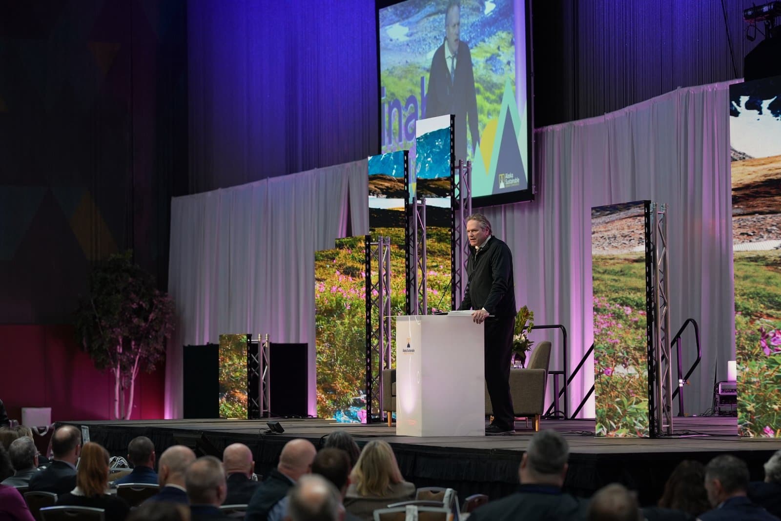 A man in a dark jacket speaks at a white podium on a stage, addressing an audience at the Anchorage Sustainable Energy Conference. Behind him, large screens display Alaskan landscapes and the 'Alaska Sustainable' logo.