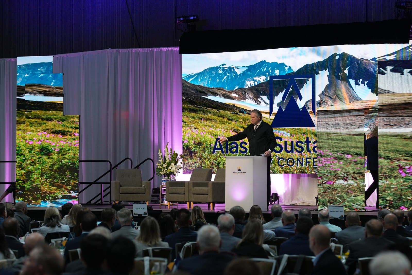 A man in a dark suit speaks from a podium on a stage, gesturing towards a large screen behind him displaying a vibrant Alaskan mountain landscape with wildflowers. An audience is seated in the foreground, facing the stage at the Alaska Sustainable Conference.