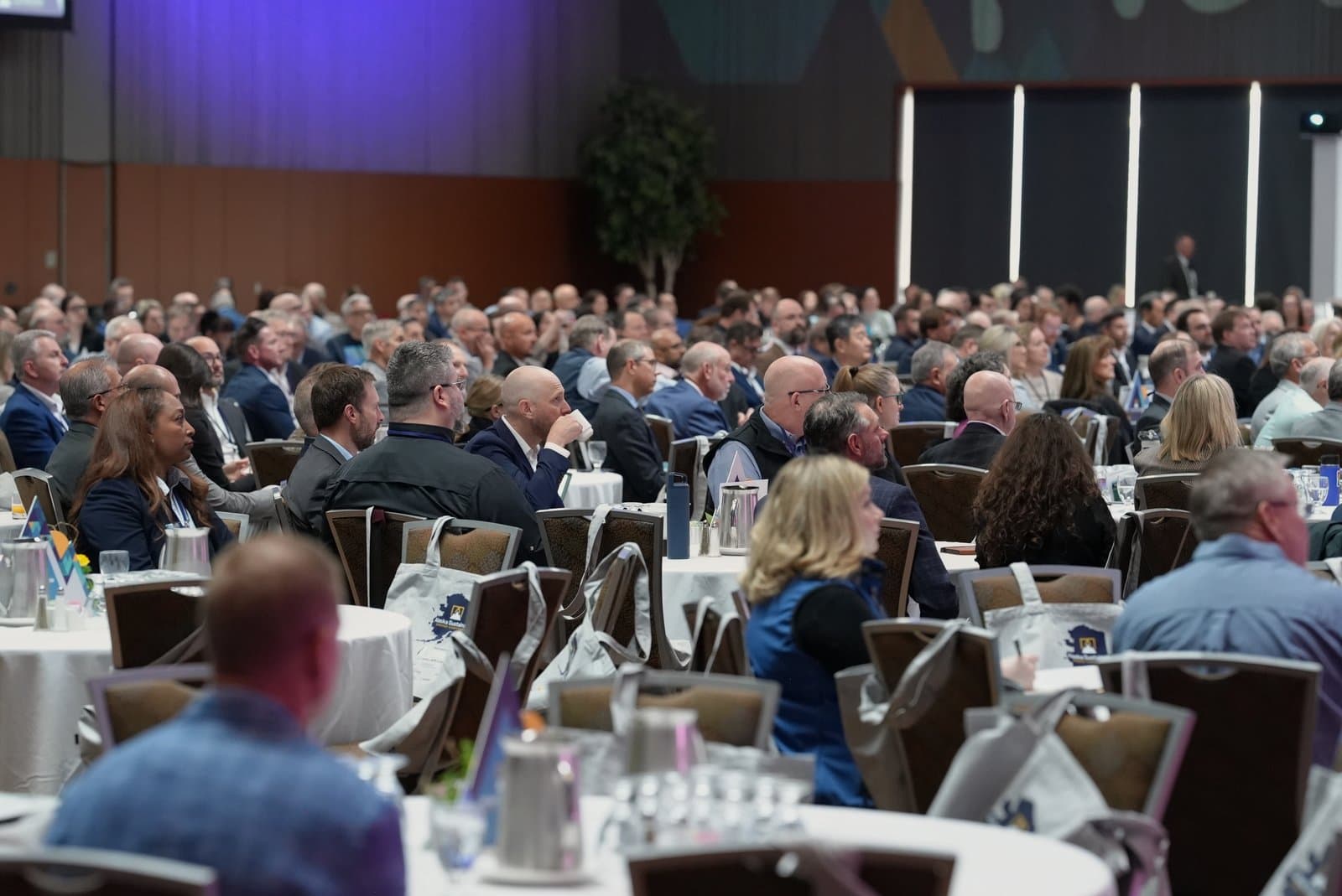 A wide shot of a large conference hall filled with numerous attendees seated at round tables, facing forward towards a stage or presentation area. Many individuals are engaged, looking ahead, while some are seen with drinks or taking notes. The room features modern lighting and a large screen in the background.