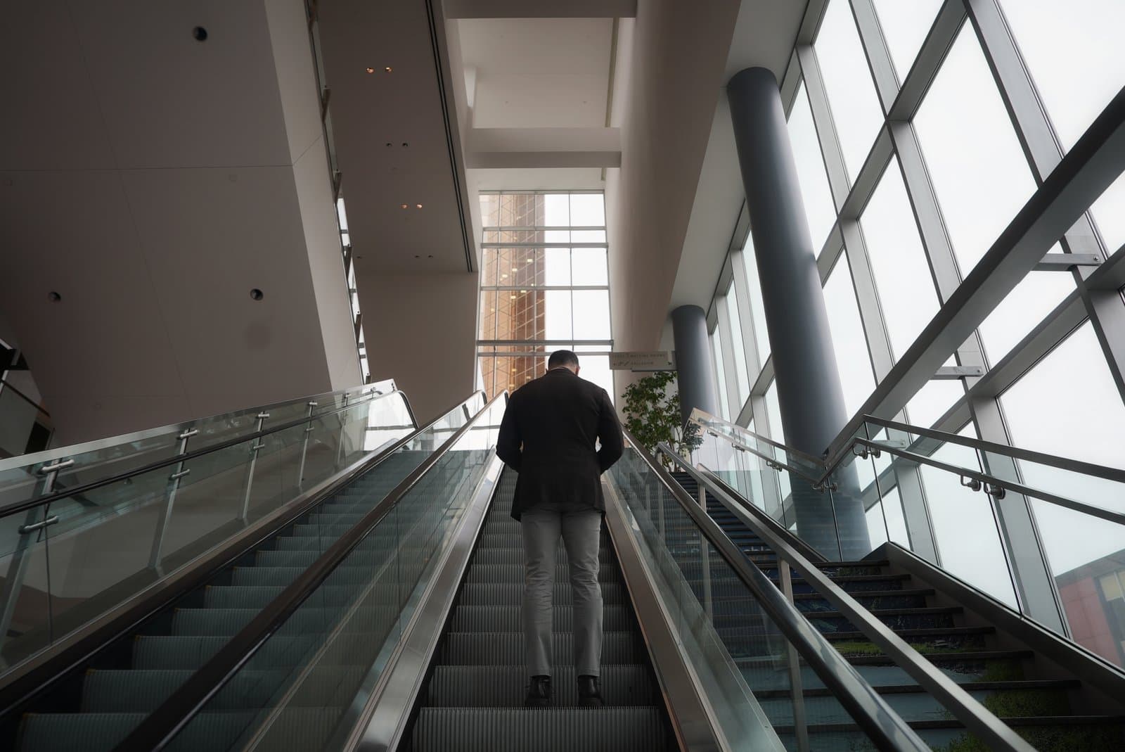 A low-angle shot from behind a person ascending an escalator in a modern building with large windows, likely at the Alaska Sustainable Energy Conference.