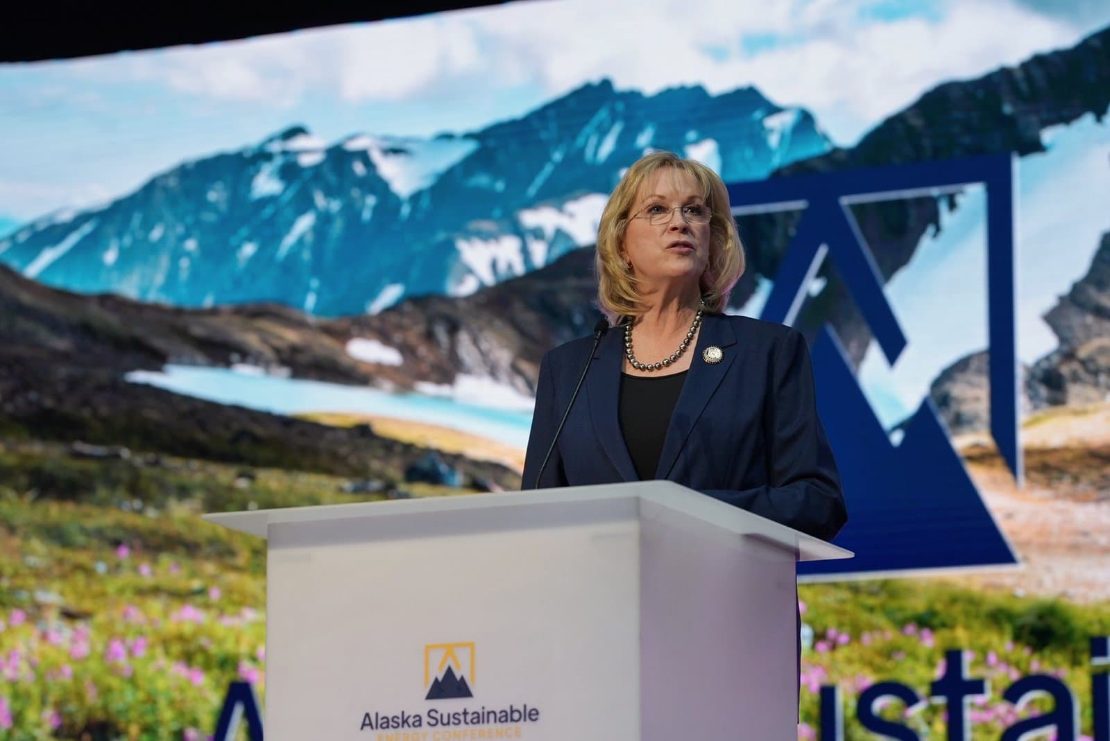 A woman with blonde hair and glasses, wearing a navy suit and pearl necklace, speaks at a white podium. Behind her, a large screen displays a scenic image of snow-capped mountains, a glacial lake, and a logo for the 'Alaska Sustainable Energy Conference'.