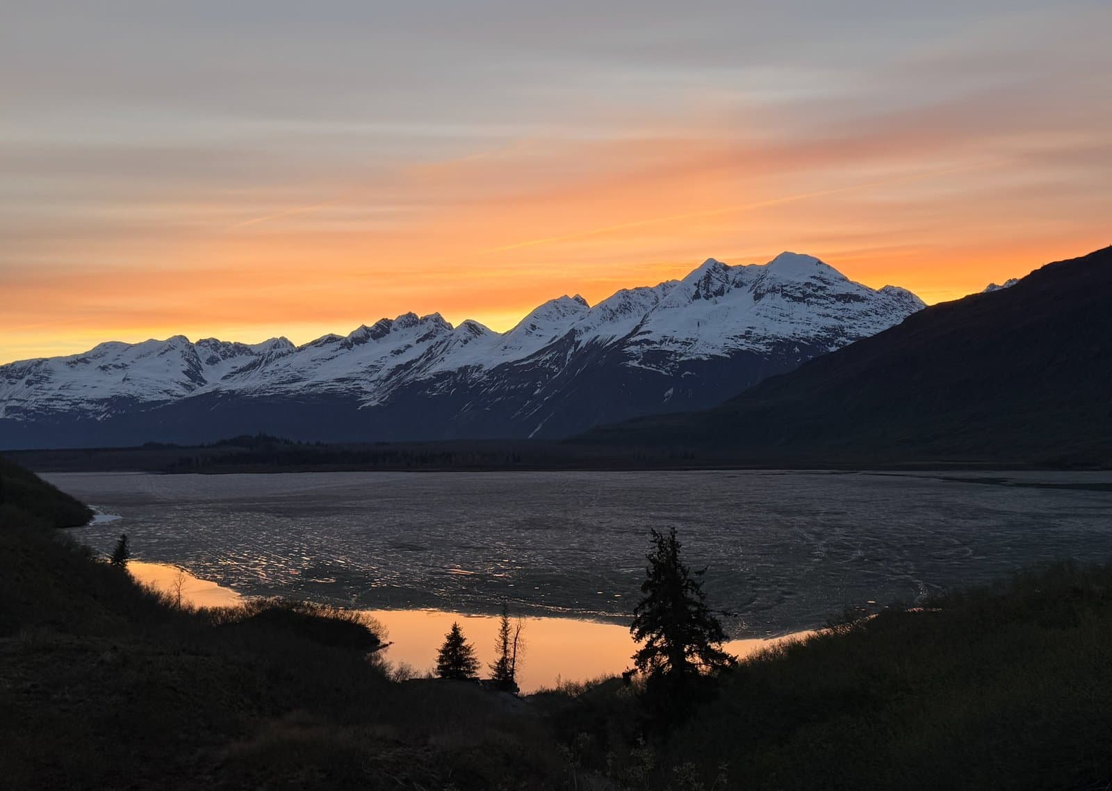 A vibrant orange sunset illuminates the snow-capped Chugach Mountains and the partially frozen waters of Robe Lake in Valdez, Alaska.