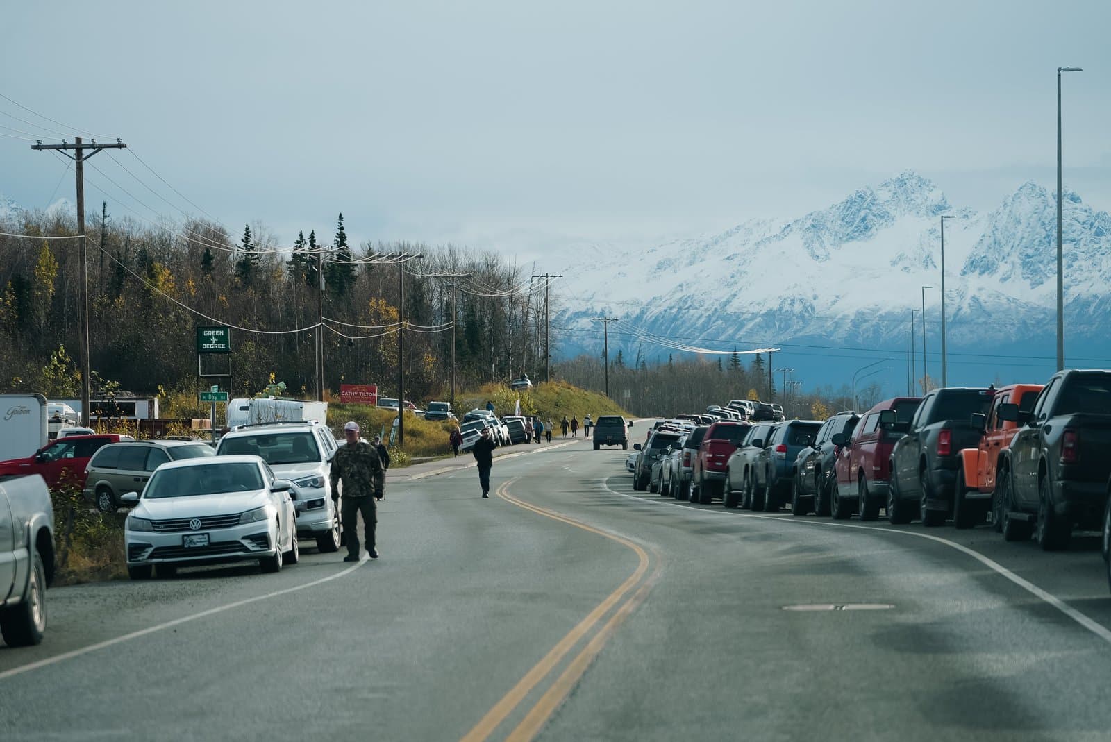 Vehicles line the shoulder of a road near a mountain range with snow-capped peaks under a clear sky in Alaska.