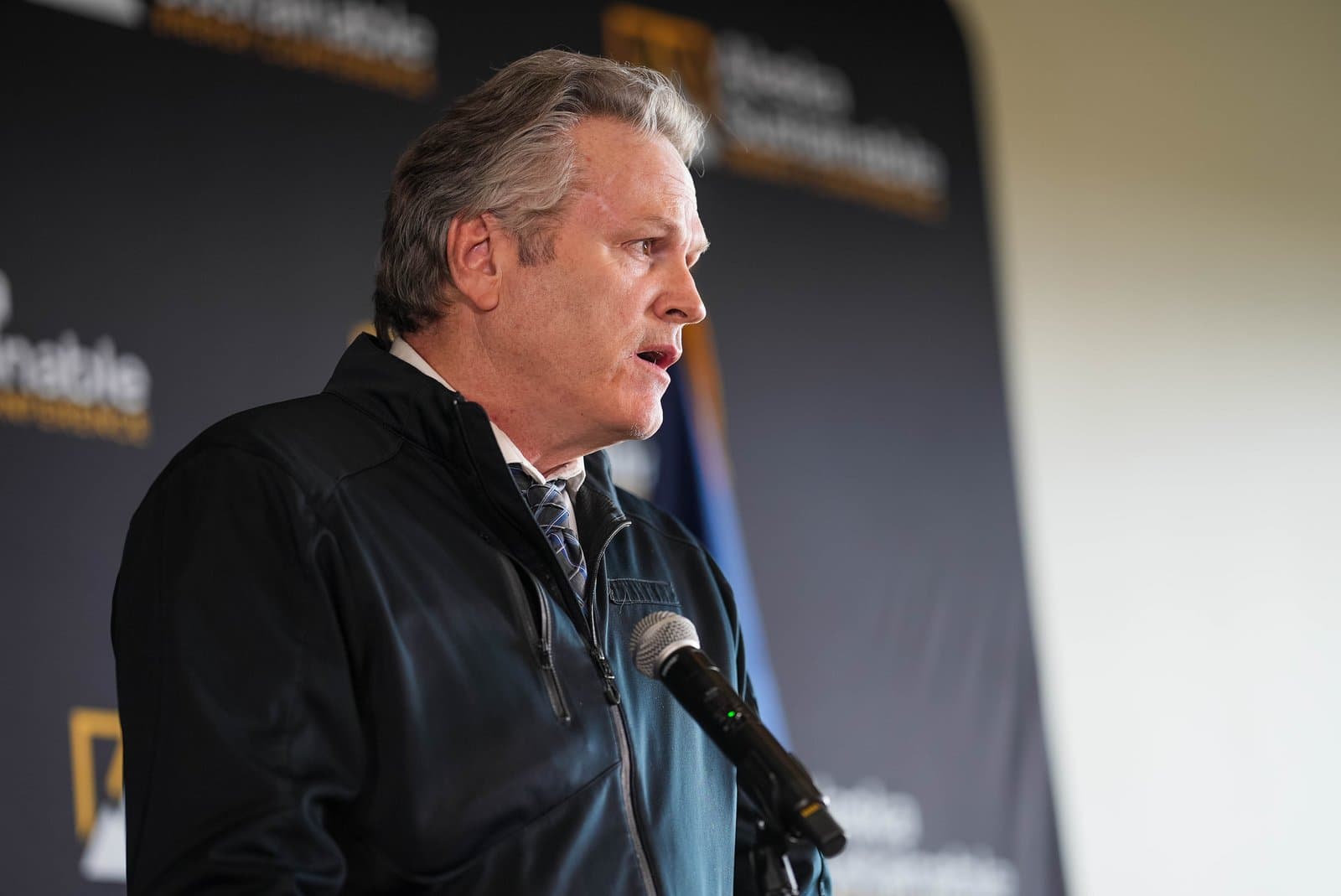 A man with gray hair, wearing a dark jacket and a patterned tie, speaks into a microphone at a podium. Behind him is a dark banner with the word "Sustainable" partially visible, indicating the Alaska Sustainable Energy Conference.