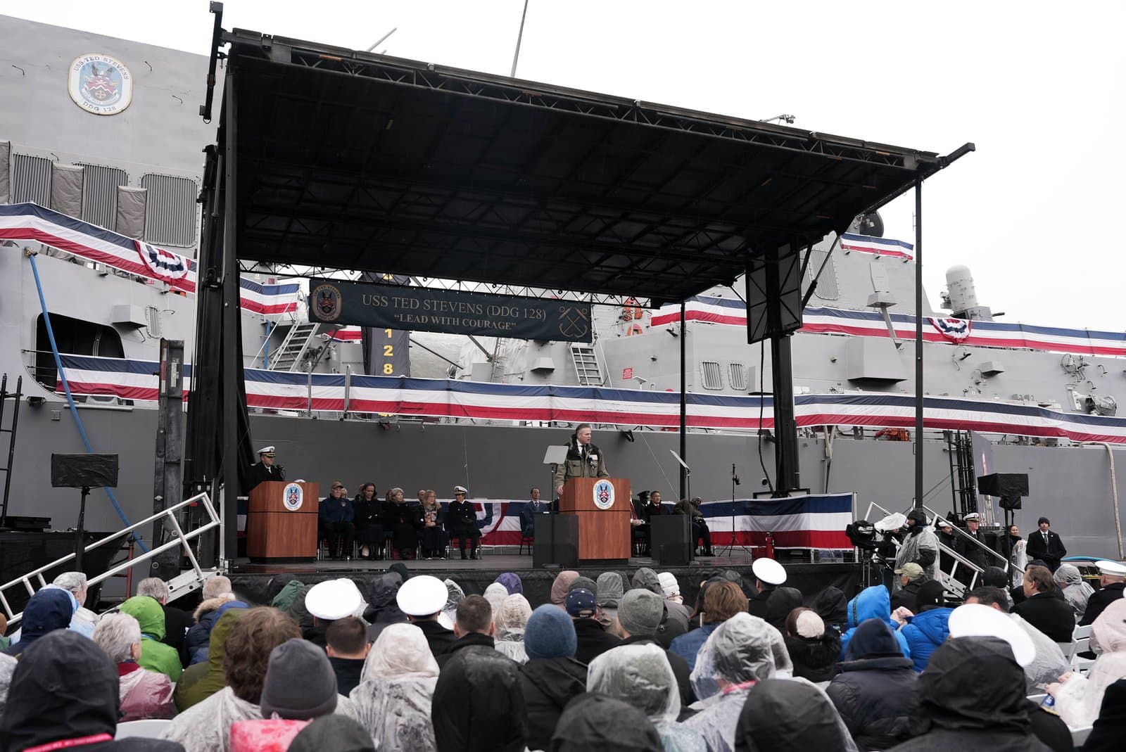 A speaker addresses a crowd from a stage in front of the USS Ted Stevens, a naval destroyer decorated with red, white, and blue bunting.