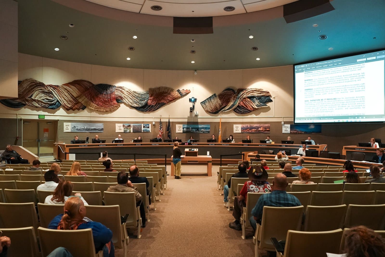 The Anchorage Assembly holds a public meeting in a circular chamber with a large projection screen displaying legislative text.