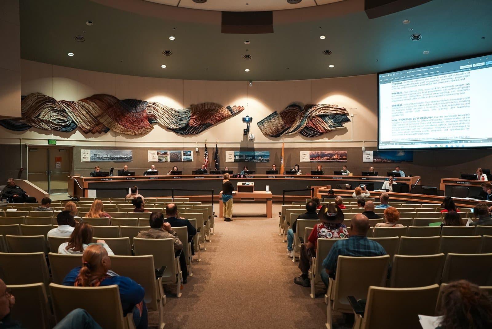 The Anchorage Assembly convenes for a public meeting with members seated at a curved dais and attendees watching from the auditorium floor.