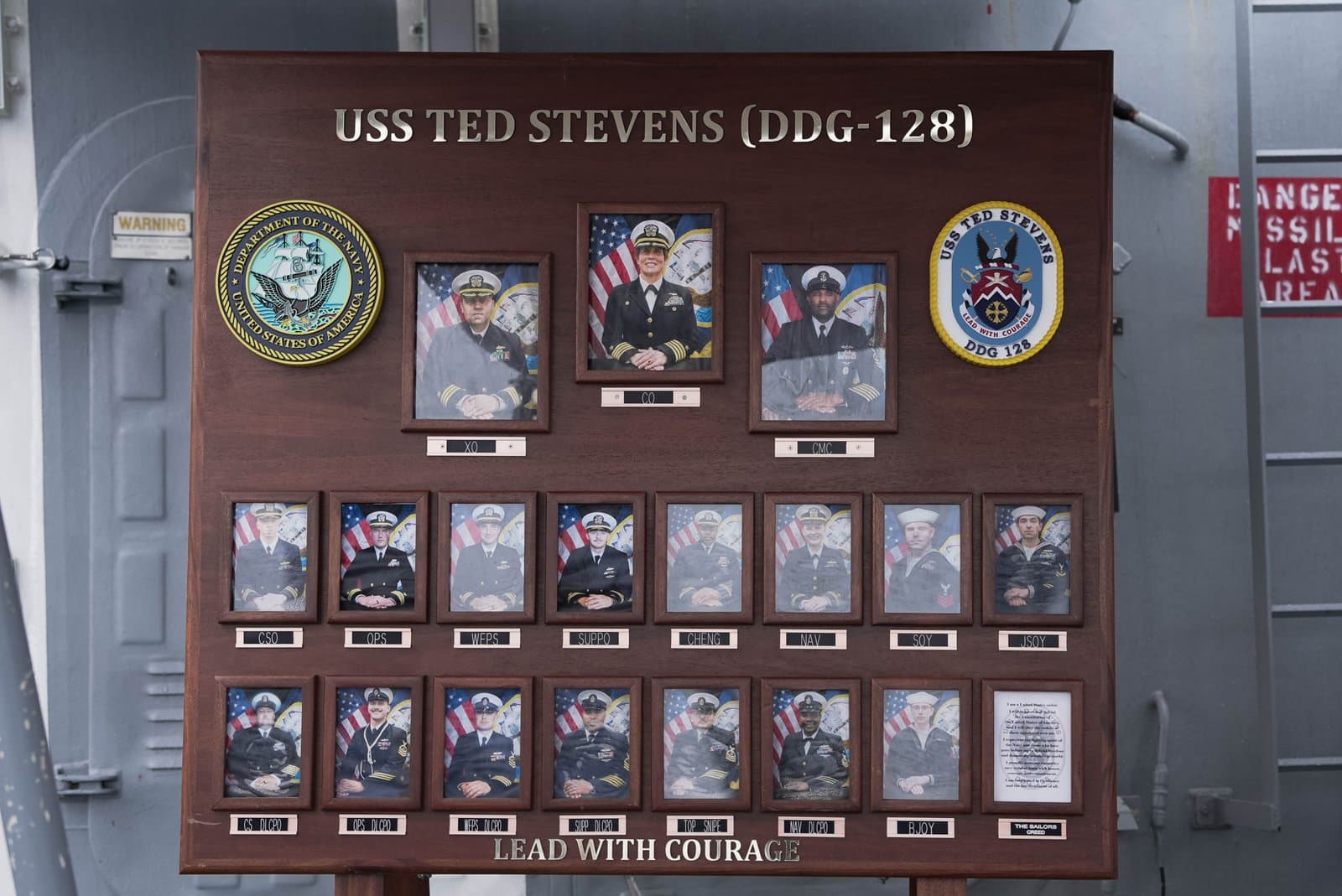 A wooden plaque on the USS Ted Stevens displays portraits of the ship's leadership and crew members alongside official Navy insignia.