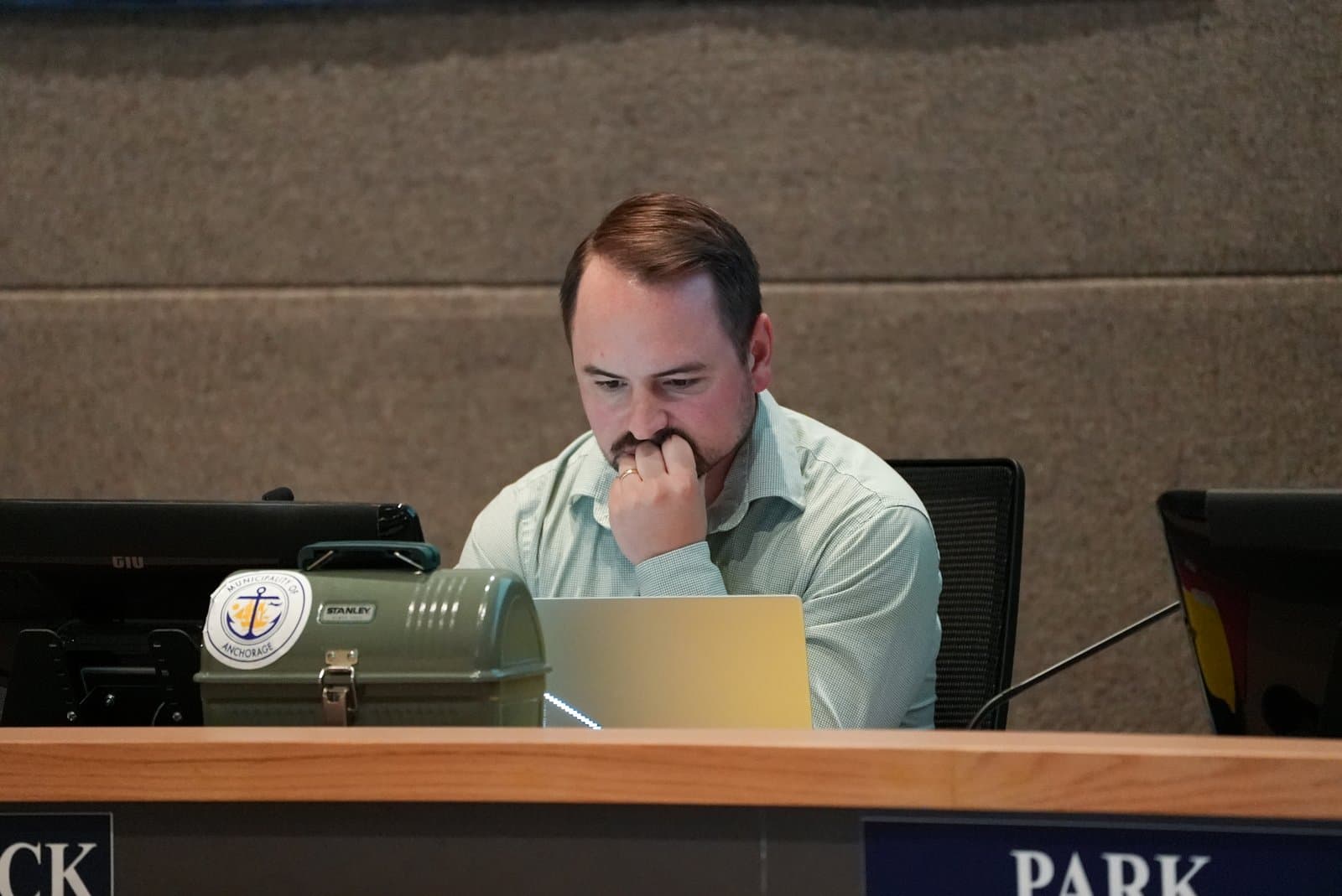 South Anchorage Assembly member Keith Mccormick at Anchorage Assembly meeting at a desk with a laptop and a Municipality of Anchorage branded lunchbox during a meeting.