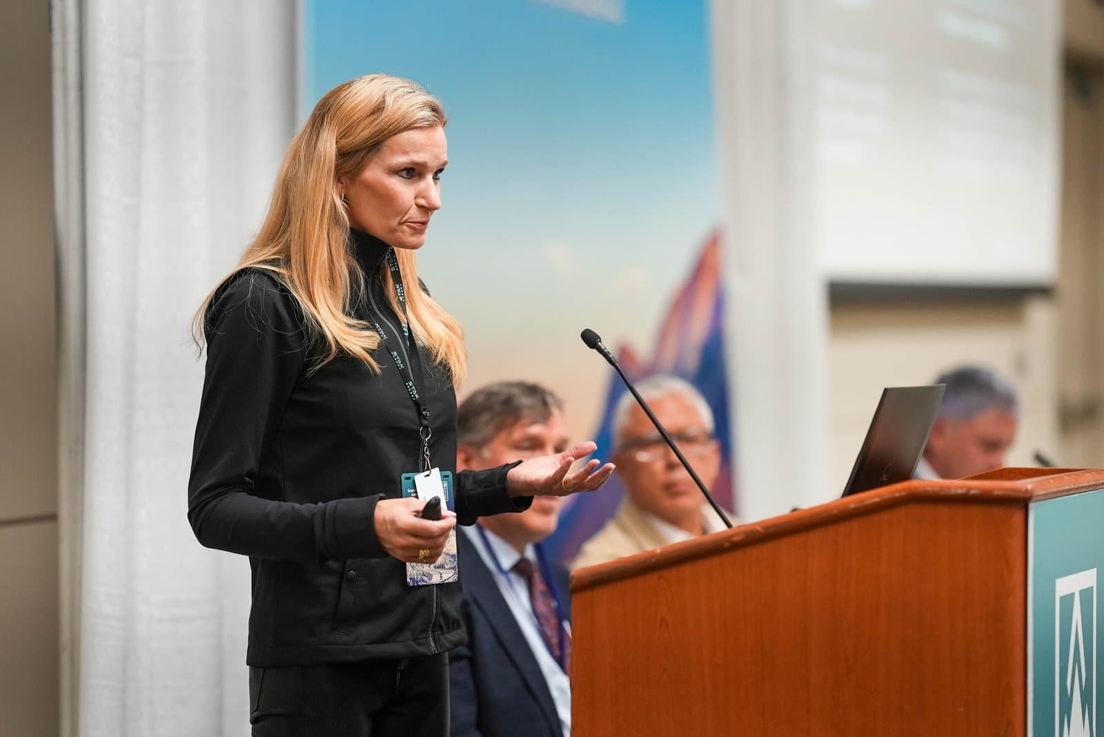 A woman with blonde hair, wearing a black jacket, speaks at a wooden podium with a microphone, gesturing with her left hand. In the background, an audience is seated, and a large banner with a blue sky and abstract shapes is visible.