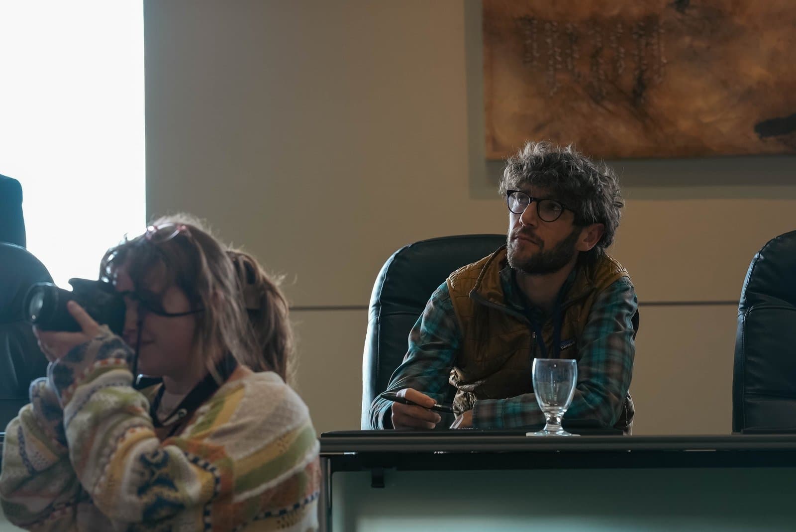 A person with a camera photographs a man seated at a table during the Alaska Sustainable Energy Conference in Anchorage. The man, wearing glasses and a vest, looks intently upwards.