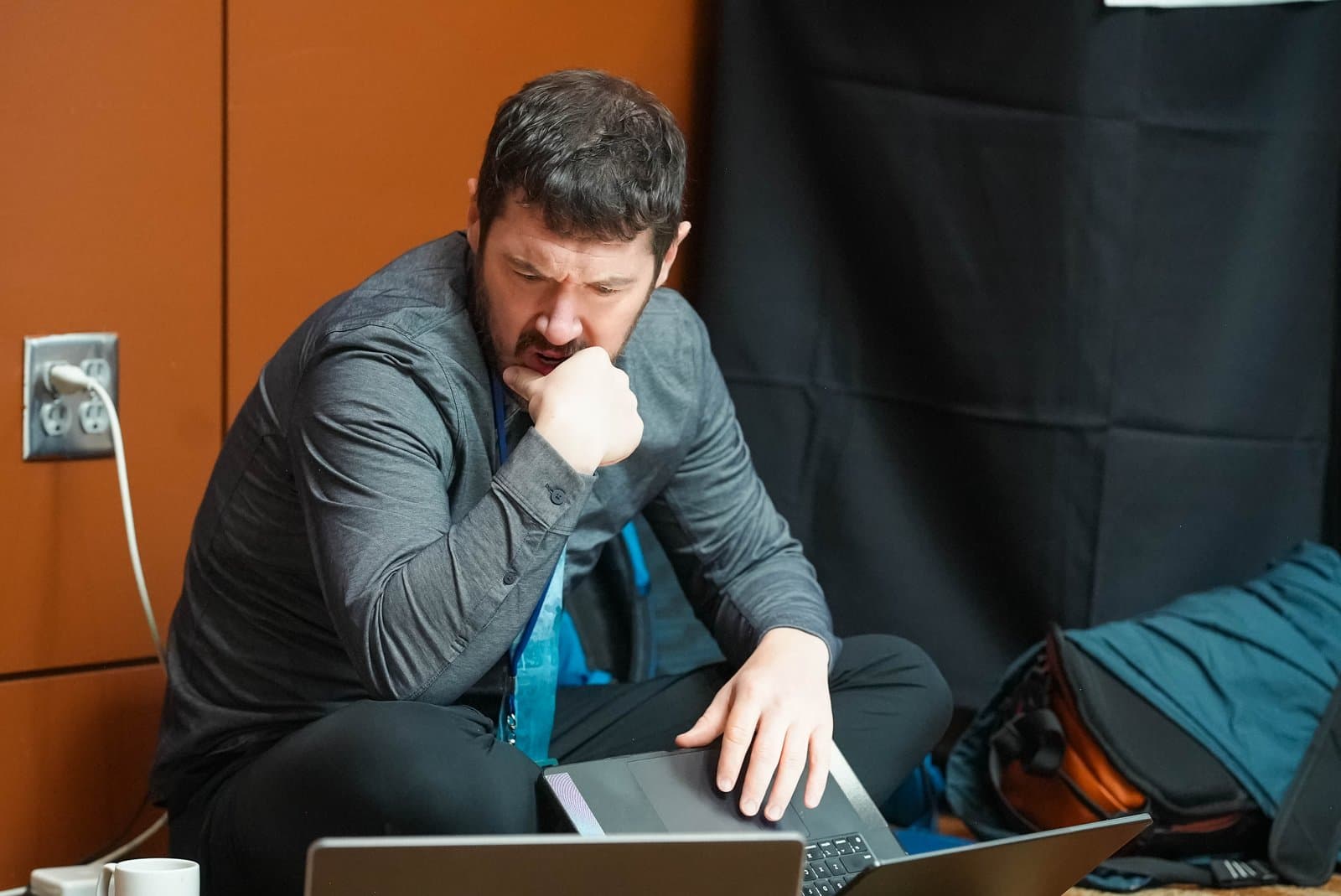A man with a beard, wearing a grey long-sleeved shirt and a blue lanyard, sits cross-legged on the floor, intently looking at a laptop with his hand on his chin, at the Alaska Sustainable Energy Conference in Anchorage.