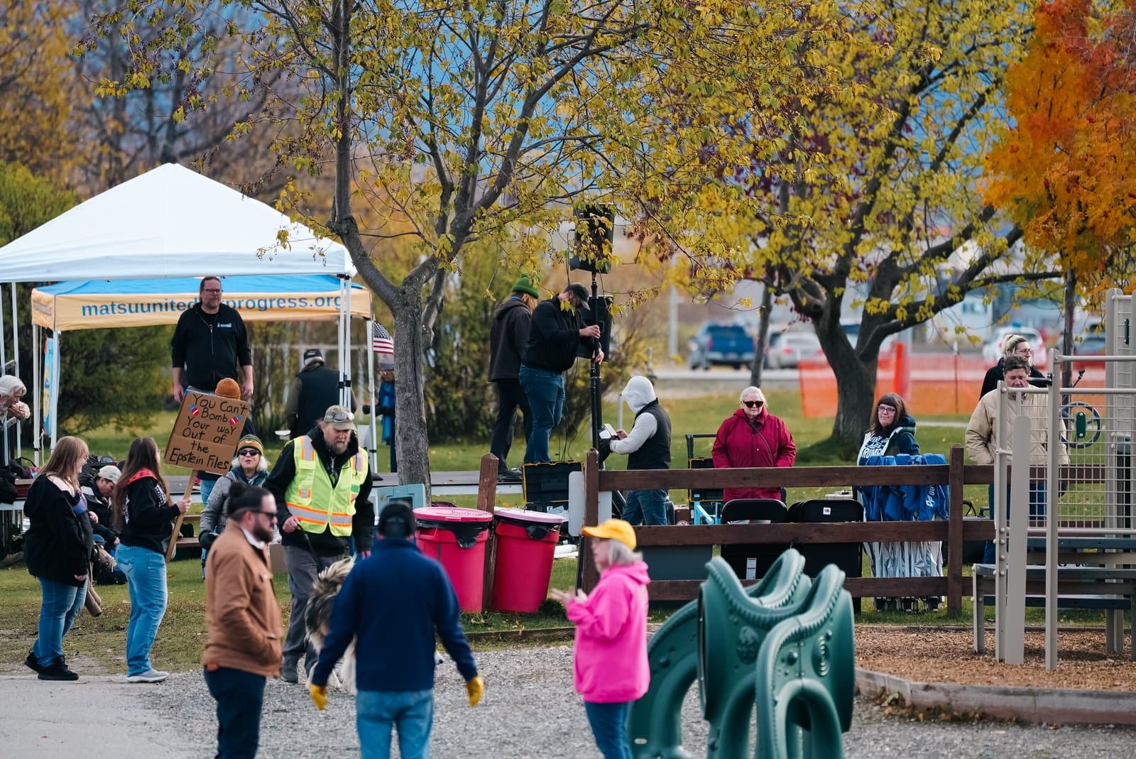 A group of people gathers in a park during an autumn day, with some individuals holding signs and a banner visible in the background.