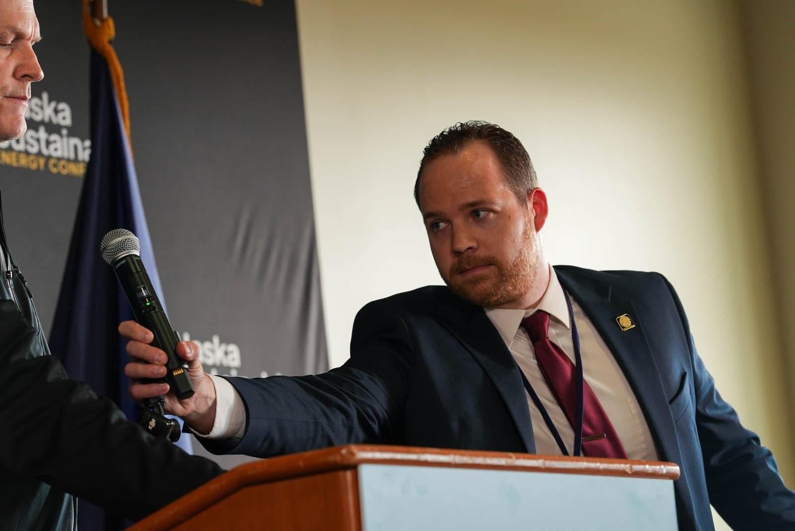 A man with a beard, wearing a dark suit and red tie, looks intently to his left while another person's hand holds a microphone in the foreground. A banner with 'Alaska Sustainable Energy Conference' is visible in the background.