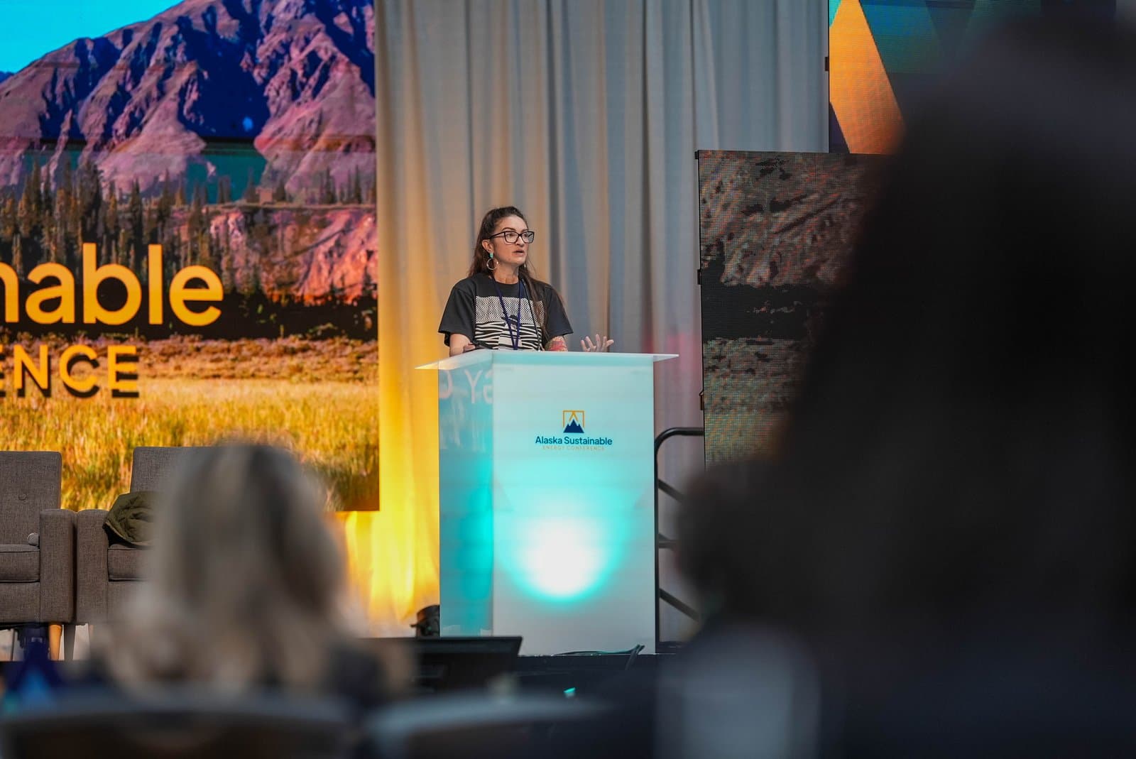 A woman with glasses and long hair speaks at a white podium with the 'Alaska Sustainable' logo. Behind her, a large screen displays a scenic Alaskan landscape and the text 'Sustainable Energy Conference', with blurred audience members in the foreground.