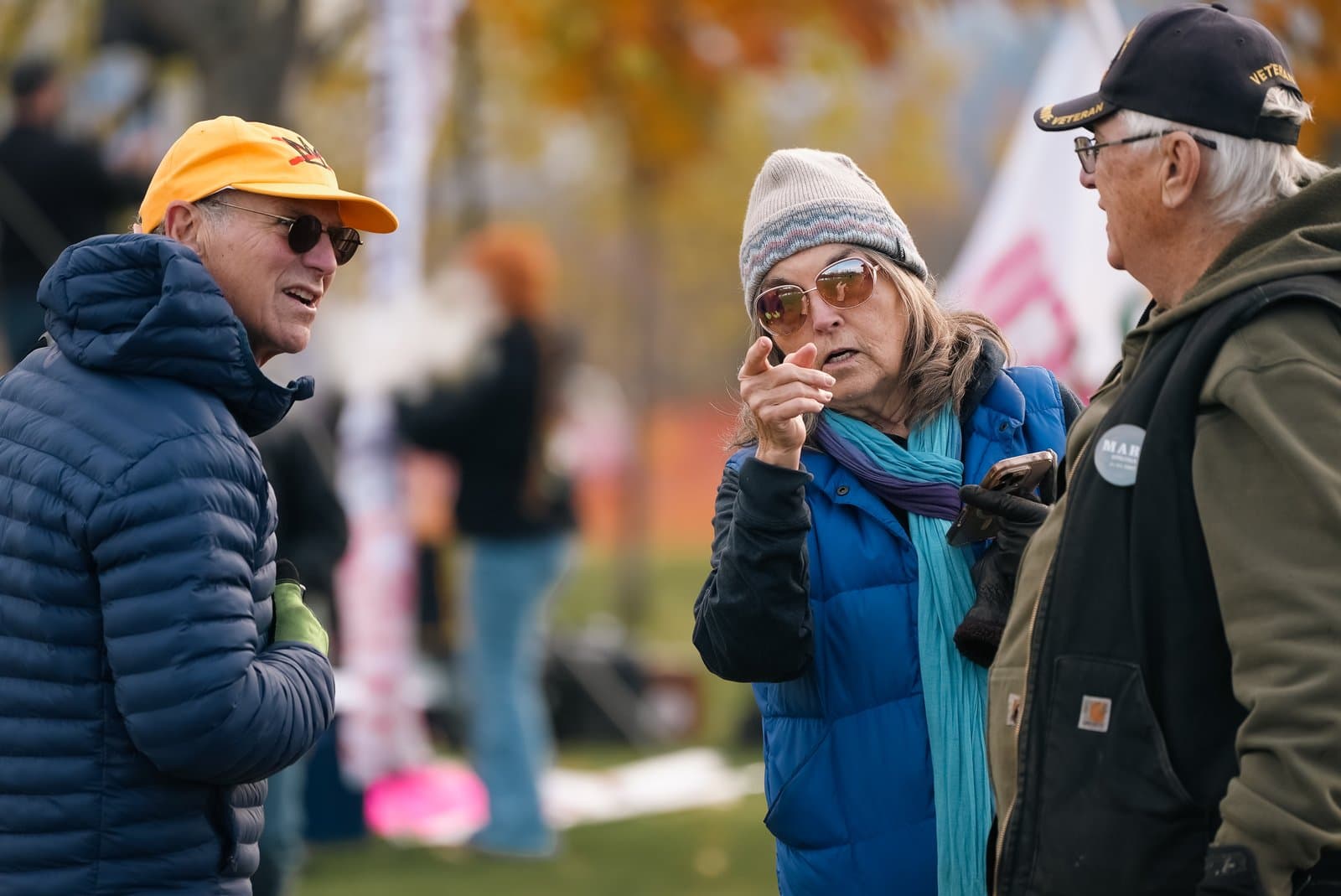 Three people in outdoor gear engage in a conversation at an outdoor community event during a crisp autumn day.