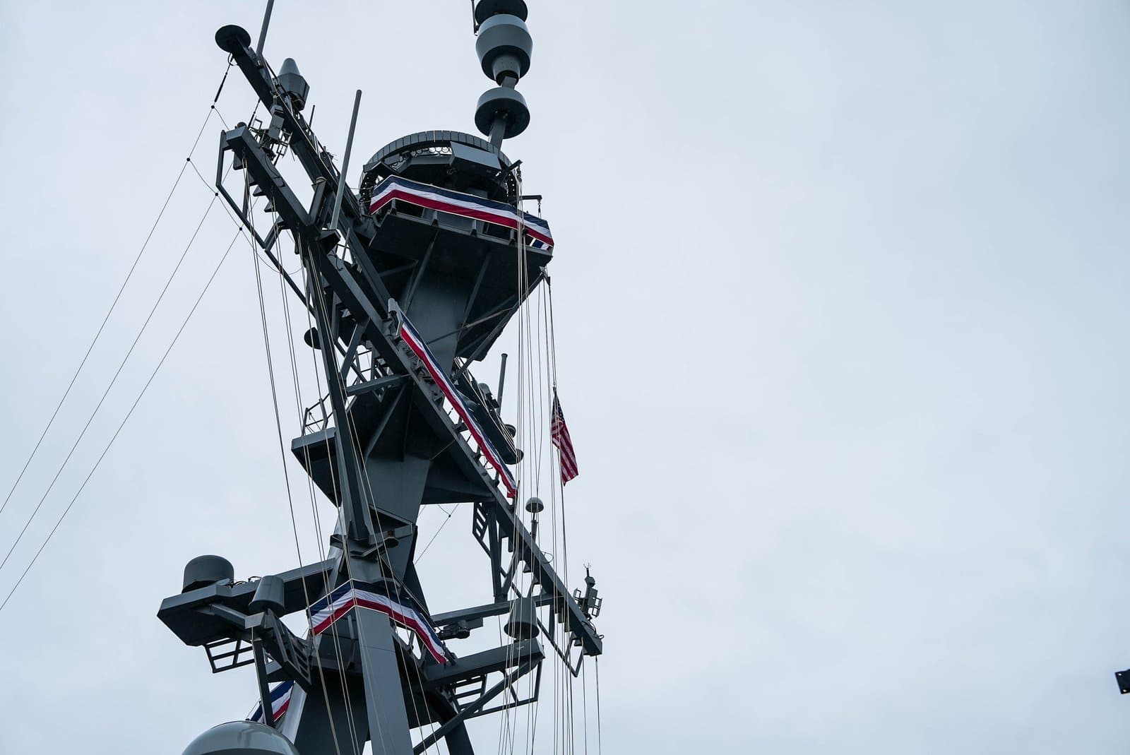 The mast of a naval vessel features radar equipment, antennas, and a small American flag flying against a cloudy, overcast sky.