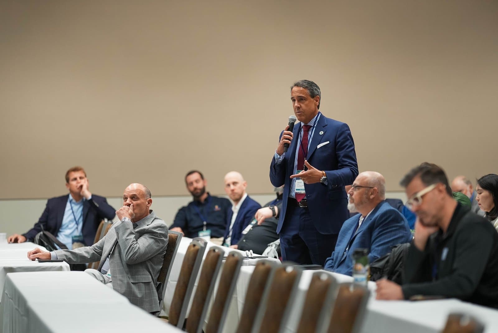 A man in a blue suit speaks into a microphone at a conference, addressing an audience of diverse attendees seated at tables.