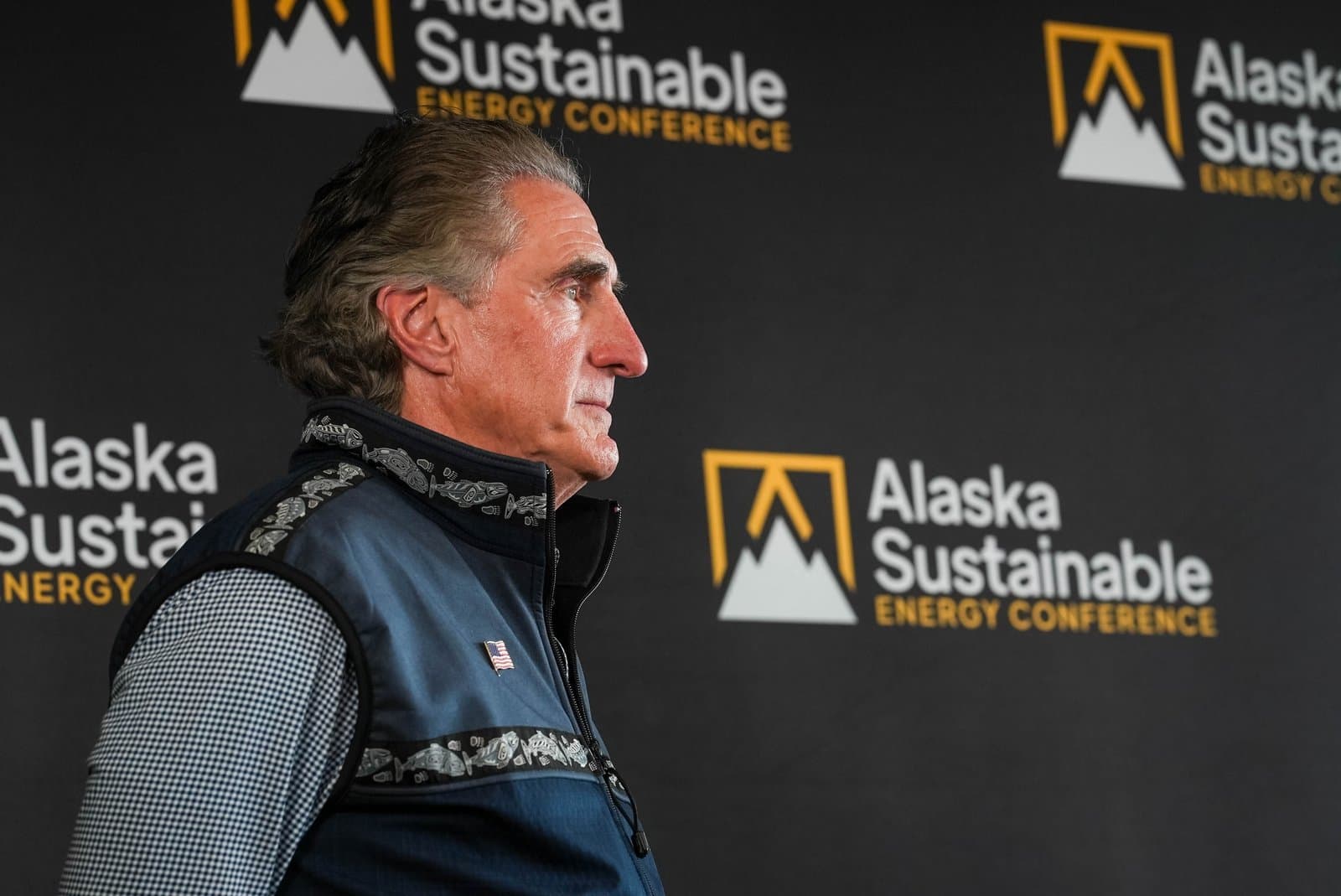 A man in a blue vest and checkered shirt is shown in profile, looking to the right, against a dark backdrop featuring the 'Alaska Sustainable Energy Conference' logo repeated.