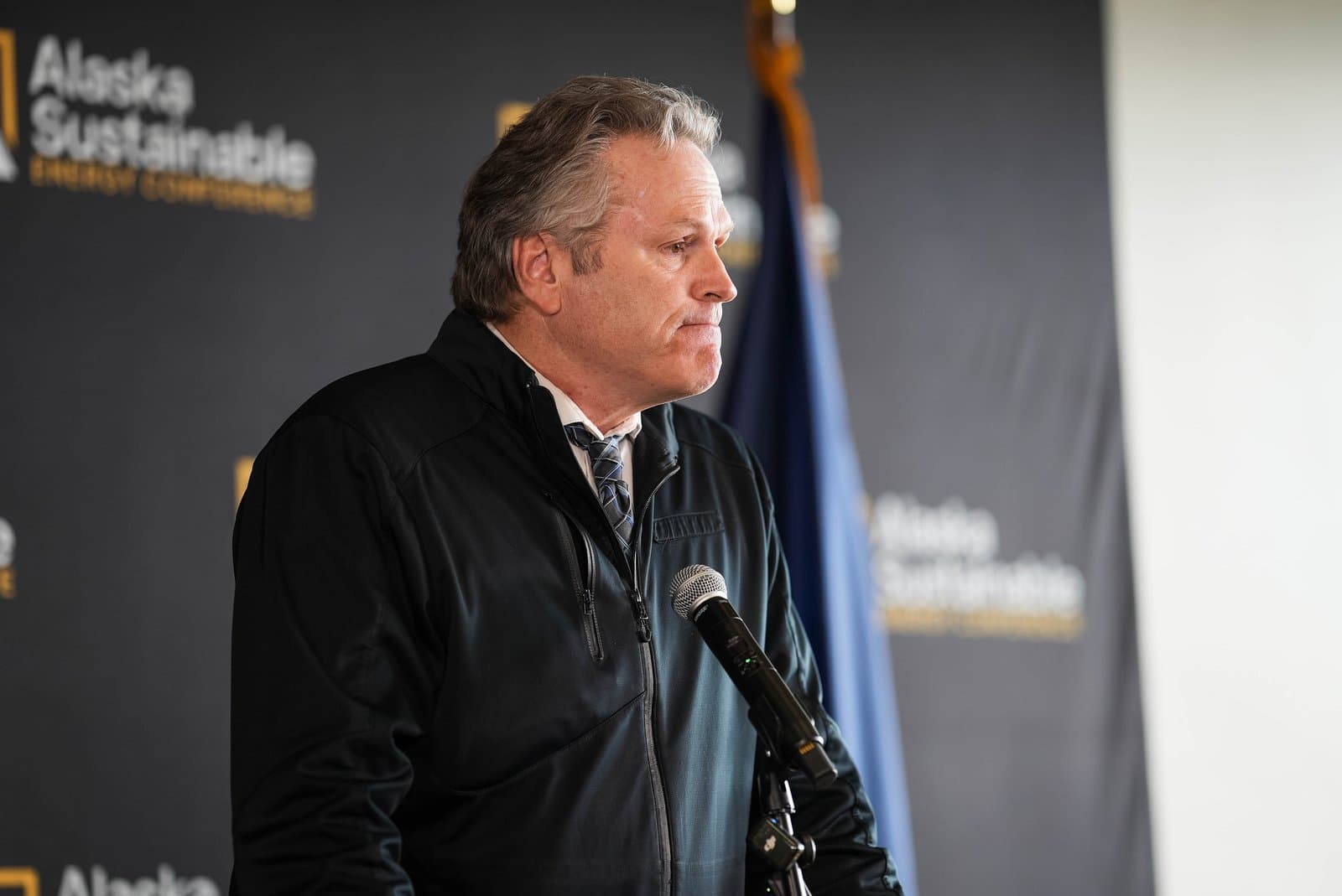 Governor Mike Dunleavy, wearing a black jacket over a white shirt and patterned tie, speaks into a microphone in profile while looking to the right. Behind him, a dark banner displays the words “Alaska Sustainable Energy Conference” in yellow text.