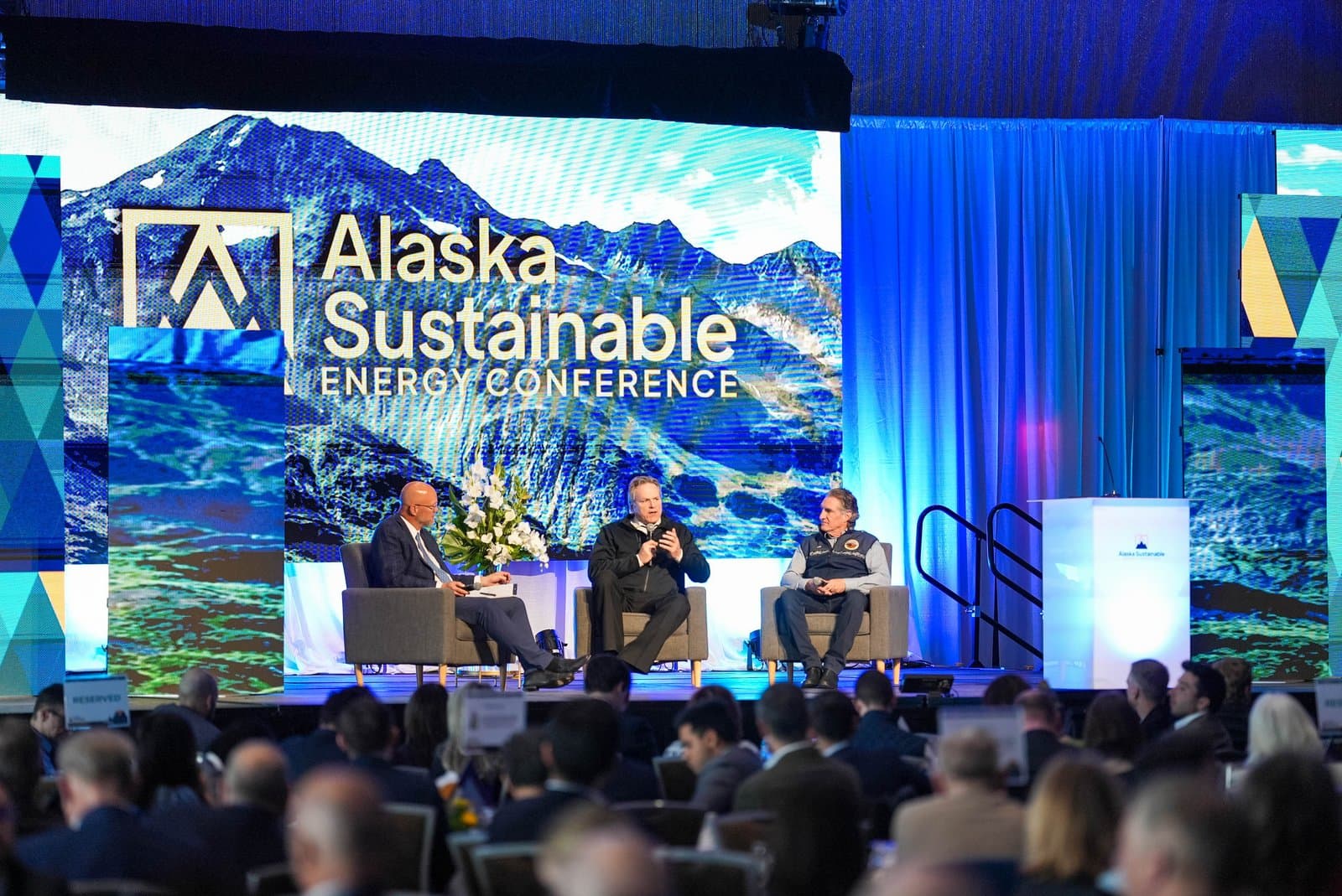 Three men are seated on a stage, speaking into microphones at the Alaska Sustainable Energy Conference. A large screen behind them displays the conference title and a scenic Alaskan landscape of mountains and water. An audience is visible in the blurred foreground.