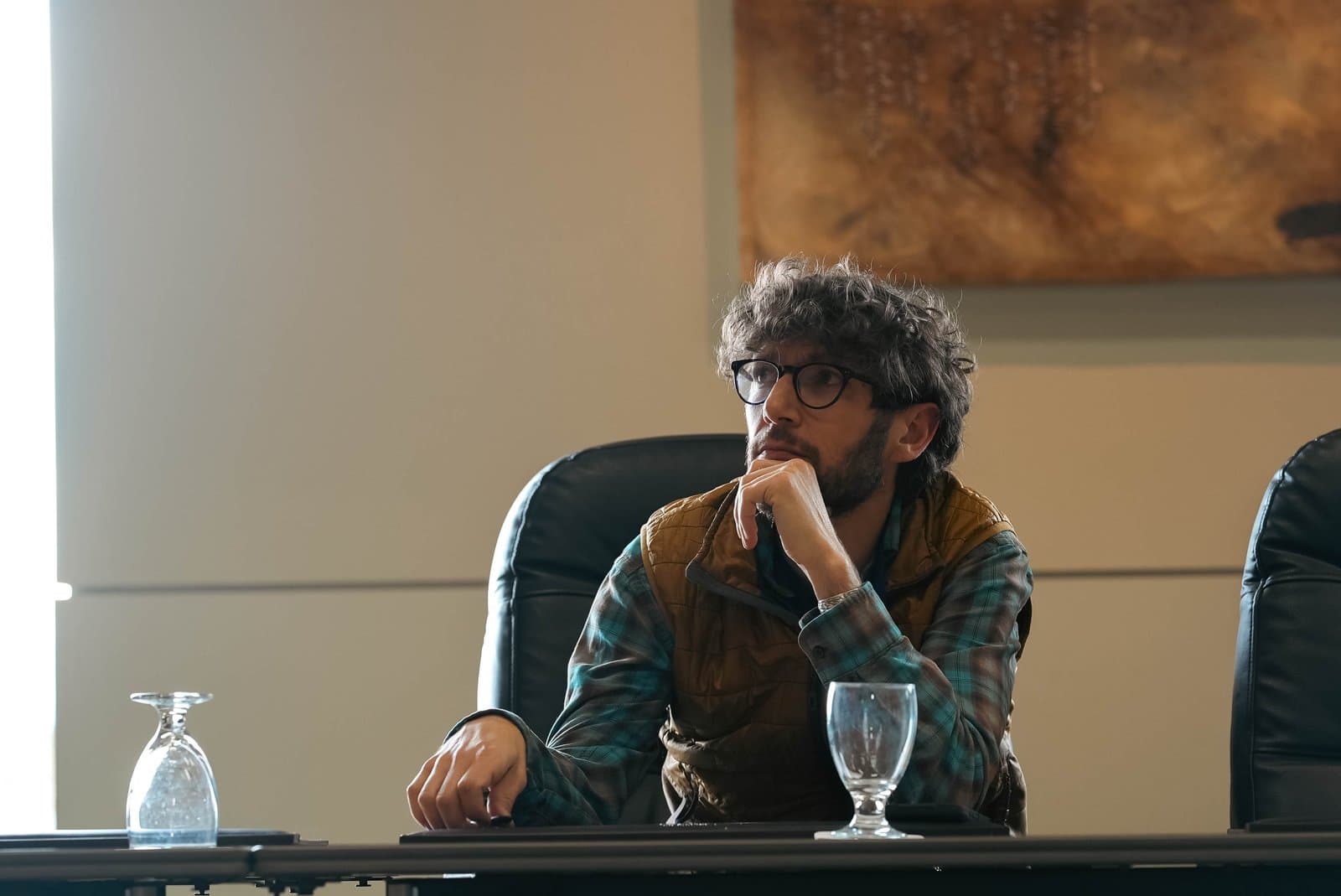 A man with curly hair and glasses, wearing a plaid shirt and brown vest, sits at a conference table, looking thoughtfully to the left with his hand on his chin. Two empty glasses are on the table in front of him, and an abstract artwork hangs on the wall in the background.
