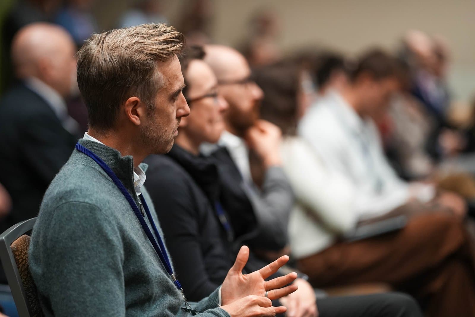 A man in a grey sweater and blue lanyard sits in profile, listening intently at an indoor conference in Anchorage. Other blurred attendees are visible in the background, suggesting a well-attended event.