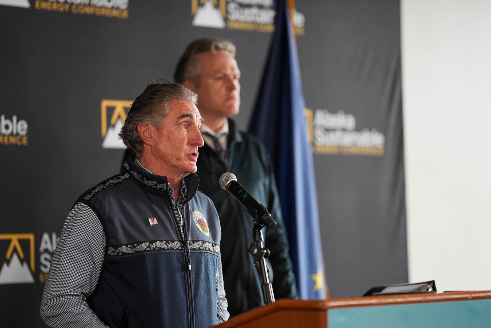 A man with gray hair and a patterned vest speaks into a microphone at a podium. Another man stands behind him, and an Alaskan flag is visible next to a backdrop that reads 'Alaska Sustainable Energy Conference'.