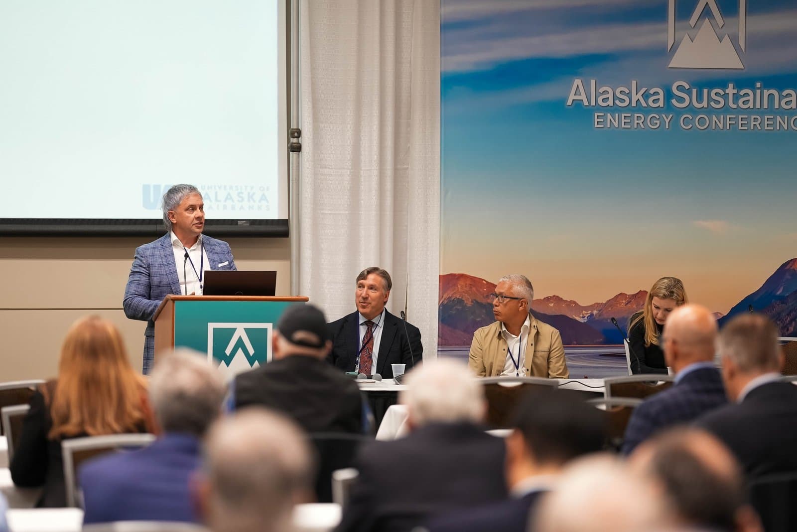 A man with gray hair speaks at a podium during the Alaska Sustainable Energy Conference, with a banner depicting mountains and water behind him, and an audience in the foreground.