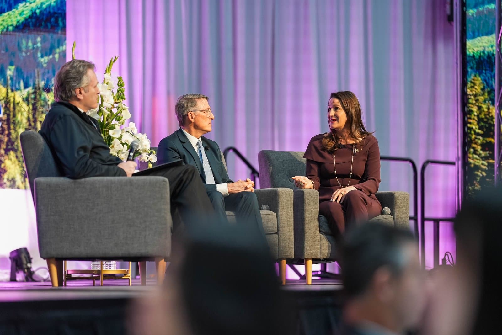 Three panelists, two men and one woman, sit on a stage in comfortable chairs, engaged in a discussion at the Alaska Sustainable Energy Conference in Anchorage. A floral arrangement is visible between two of the speakers, and the stage is backed by purple drapes and a large screen displaying a natural landscape.