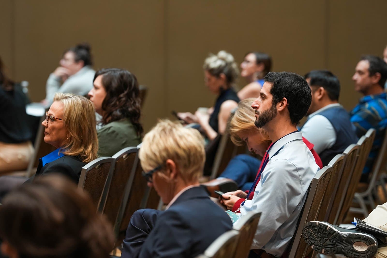A side-profile view of a diverse group of people seated in rows, attentively listening or looking at their phones during a conference. The setting is an indoor event, likely the Alaska Sustainable Energy Conference in Anchorage.