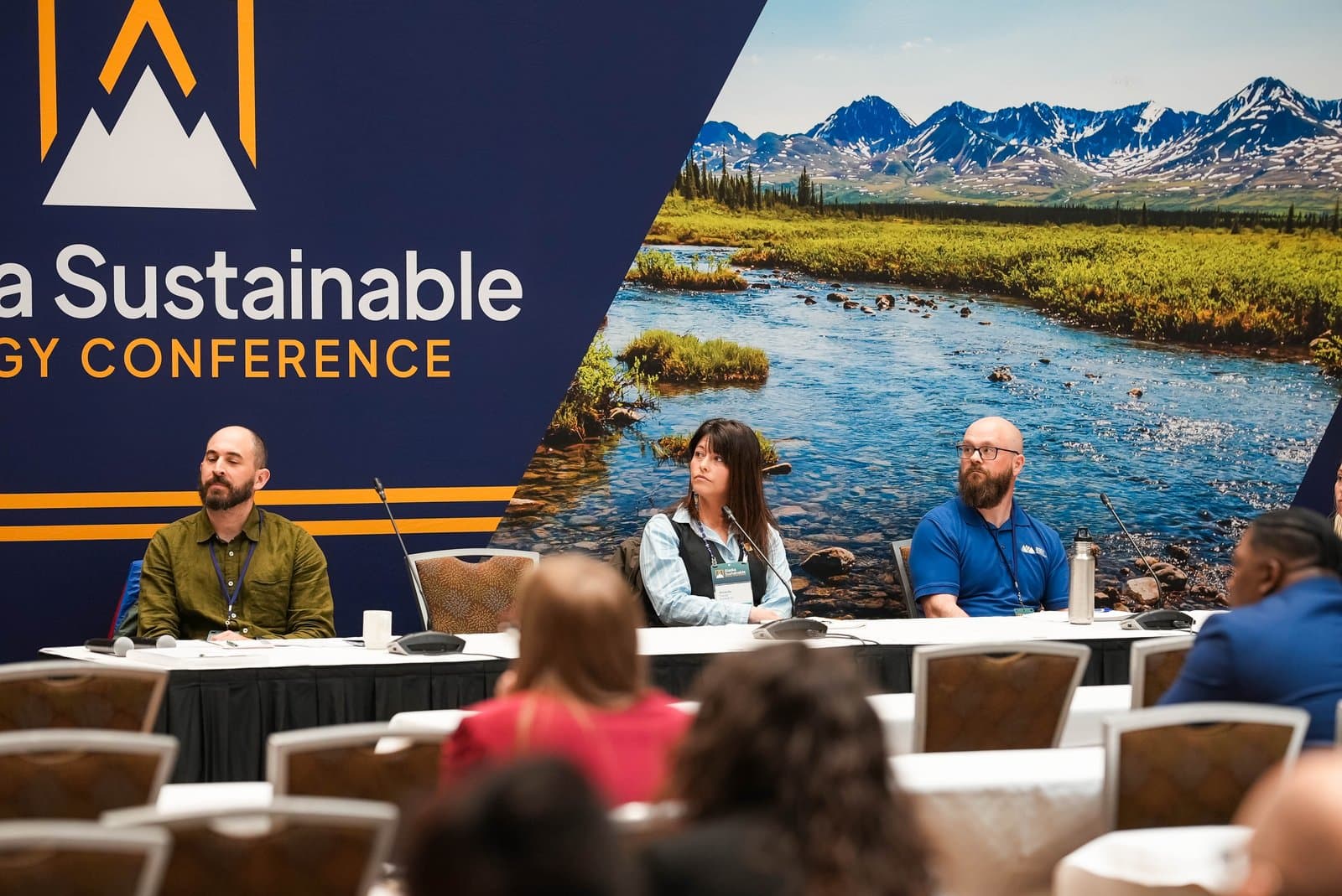 Three panelists, two men and one woman, sit at a table facing an audience during the Alaska Sustainable Energy Conference in Anchorage, with a large backdrop depicting a scenic Alaskan river and snow-capped mountains.