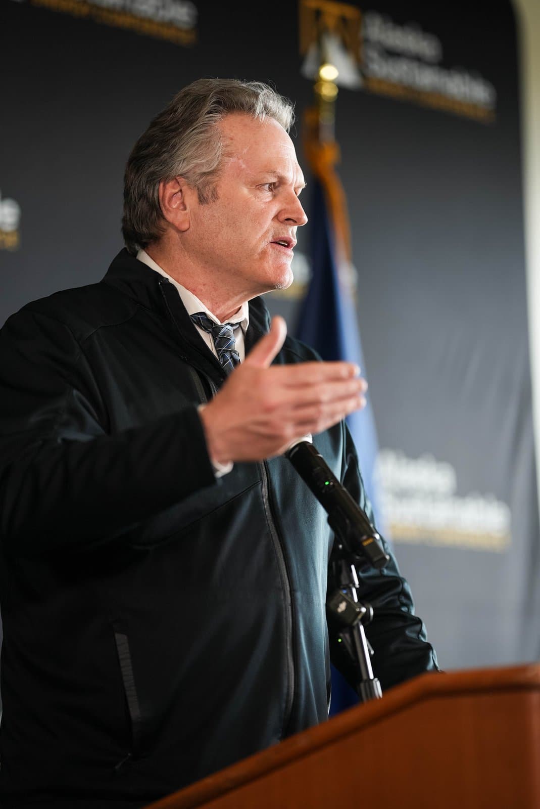 A man with grey hair, wearing a dark jacket, white shirt, and patterned tie, speaks at a podium with a microphone, gesturing with his right hand. A banner for the 'Alaska Sustainable Energy Conference' is partially visible in the background.
