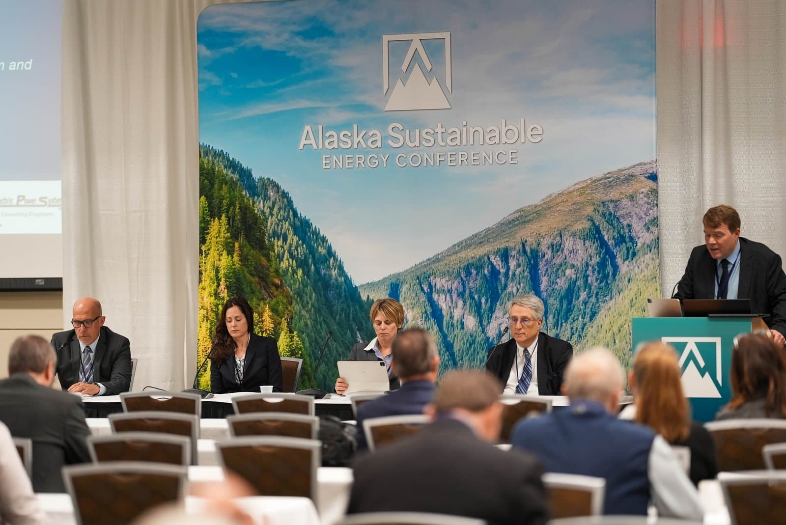 A panel of five individuals, four seated at a table and one standing at a podium, participate in the Alaska Sustainable Energy Conference. A large backdrop featuring a mountain landscape and the conference title is visible behind them.
