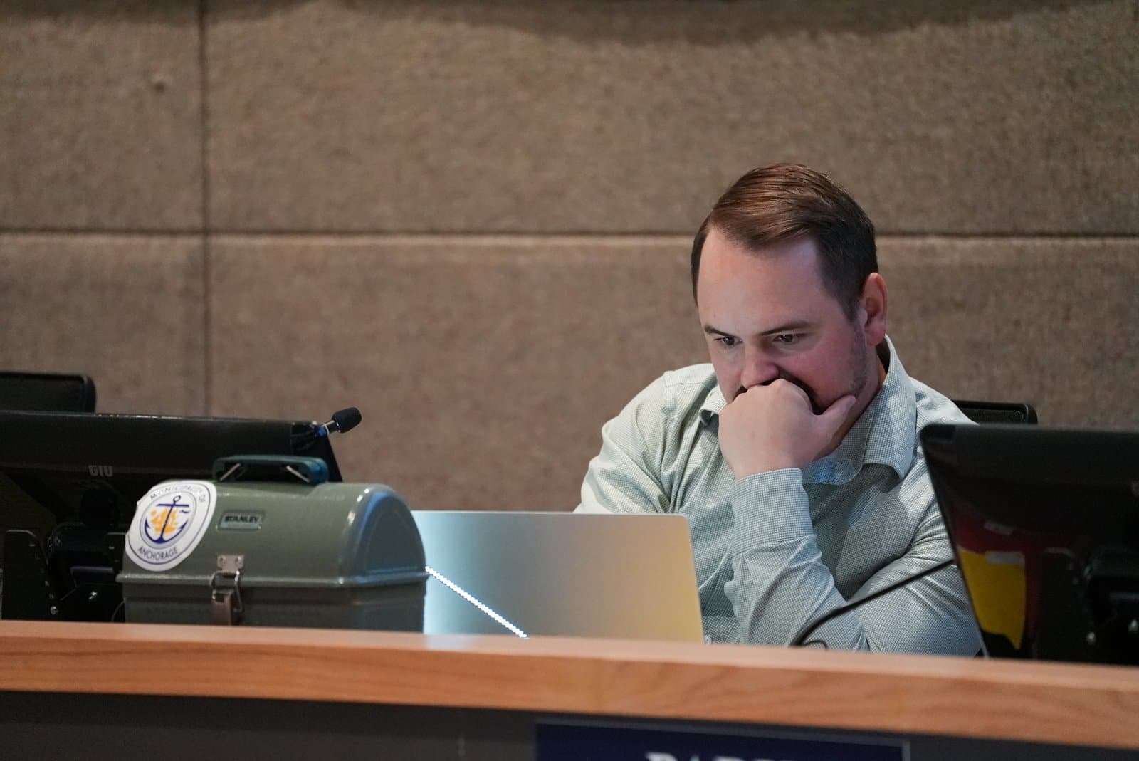 An Anchorage Assembly member sits at a desk with a laptop and a Municipality of Anchorage lunchbox during a public meeting.
