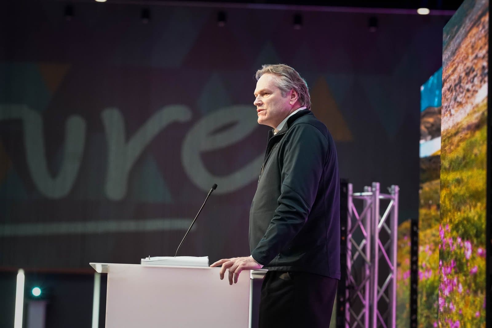 A man with gray hair, dressed in a dark jacket and white shirt, stands at a white podium with a microphone, addressing an audience at an indoor conference. Behind him, a large screen displays text, and another shows a vibrant natural landscape.