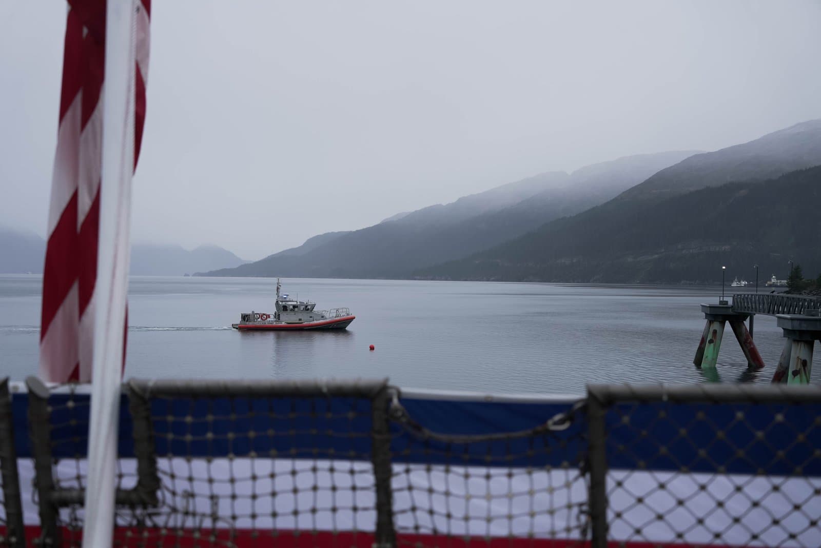 A United States Coast Guard boat patrols calm, misty waters near a pier in a mountainous coastal region during an overcast day.