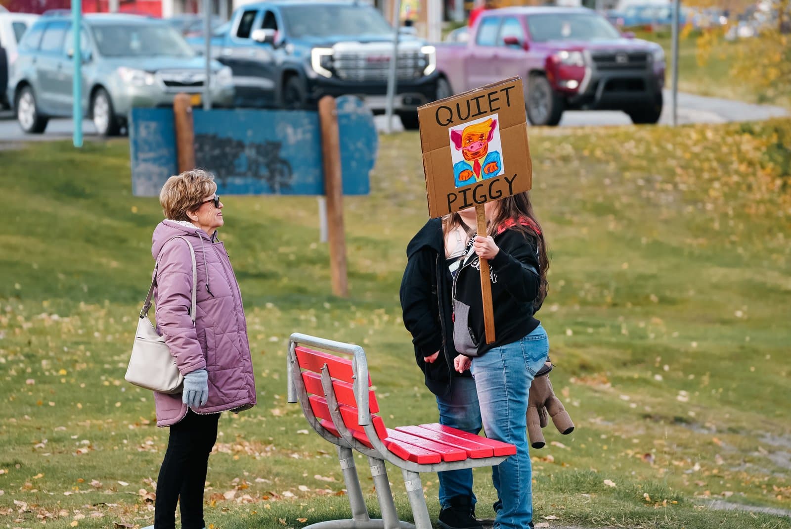 Two individuals stand near a red park bench holding a protest sign depicting a caricature of Donald Trump labeled Quiet Piggy.