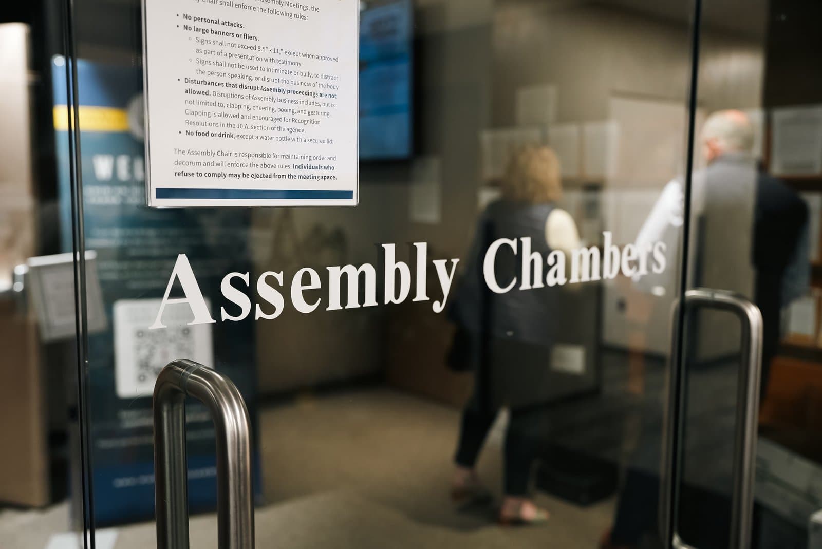 A glass door marked Assembly Chambers features a posted list of meeting rules at the entrance to an Anchorage government meeting space.
