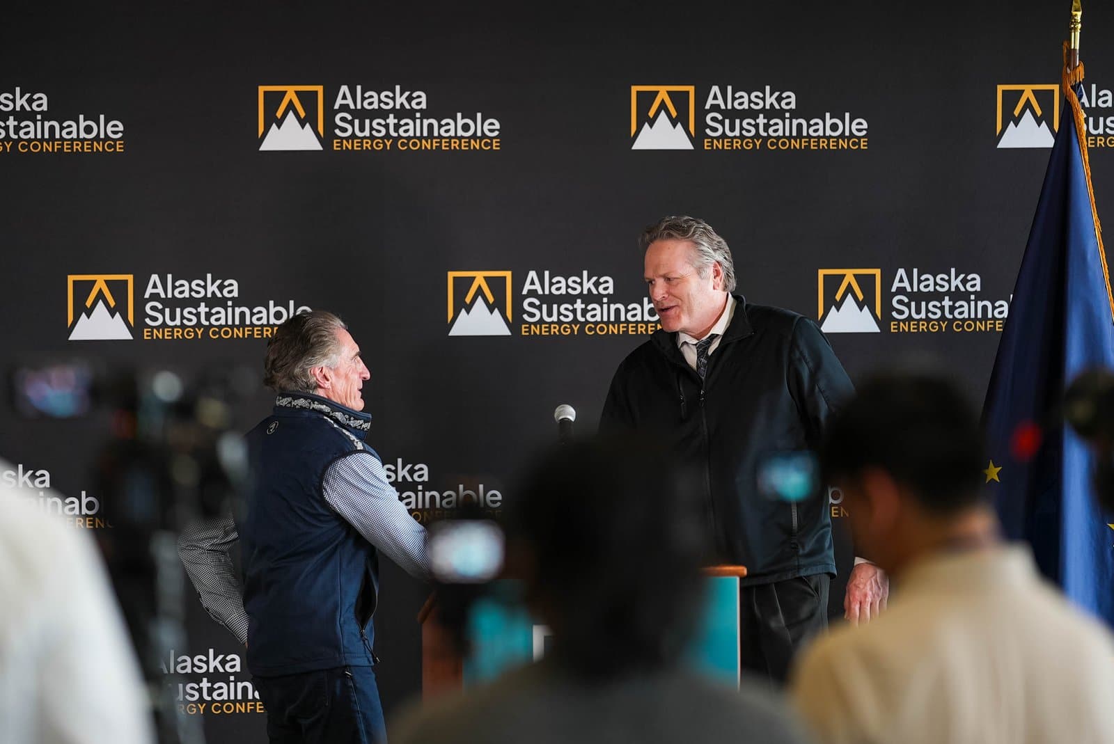 Two men are engaged in conversation at a podium during the Alaska Sustainable Energy Conference in Anchorage. The backdrop features the conference logo, and an Alaskan flag is visible on the right side of the frame.