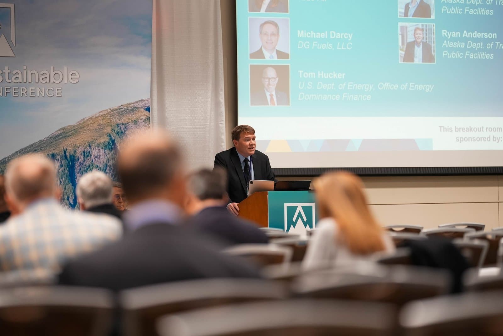 A speaker addresses an audience from a podium at the Alaska Sustainable Energy Conference in Anchorage, with a large screen displaying speaker names and affiliations, and a mountain landscape backdrop.