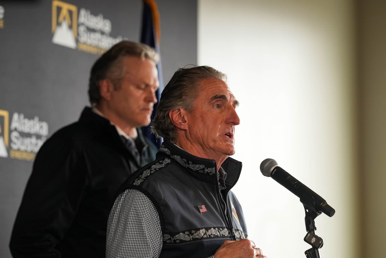 A man with grey hair wearing a dark patterned vest and an American flag pin speaks into a microphone at a podium. Another man stands slightly behind him to the left. A banner in the background reads 'Alaska Sustain' with a mountain logo.