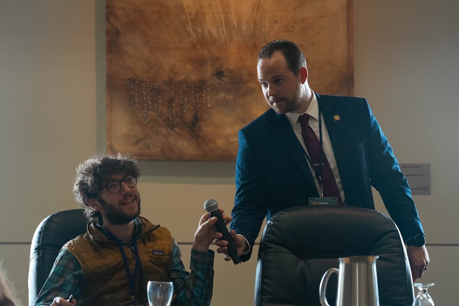 Two men, one seated and one standing, engage in a discussion during a conference. The standing man in a suit holds a microphone towards the seated man in glasses and a vest, who is smiling.
