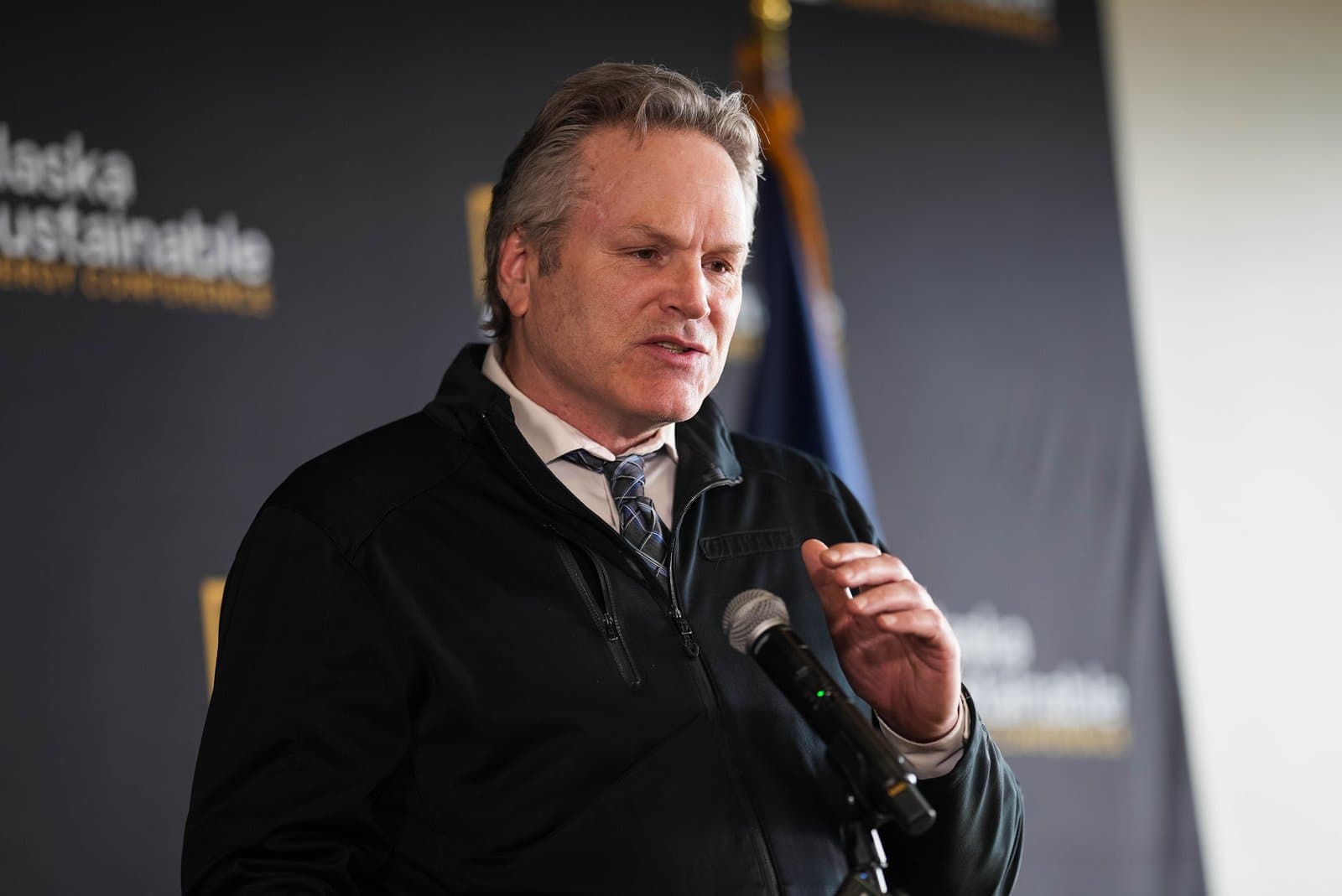 A man with grey hair, wearing a black zip-up jacket and a patterned tie, speaks into a microphone at an indoor event. A banner in the background reads 'Alaska Sustainable Energy Conference', with a partial view of a flag.