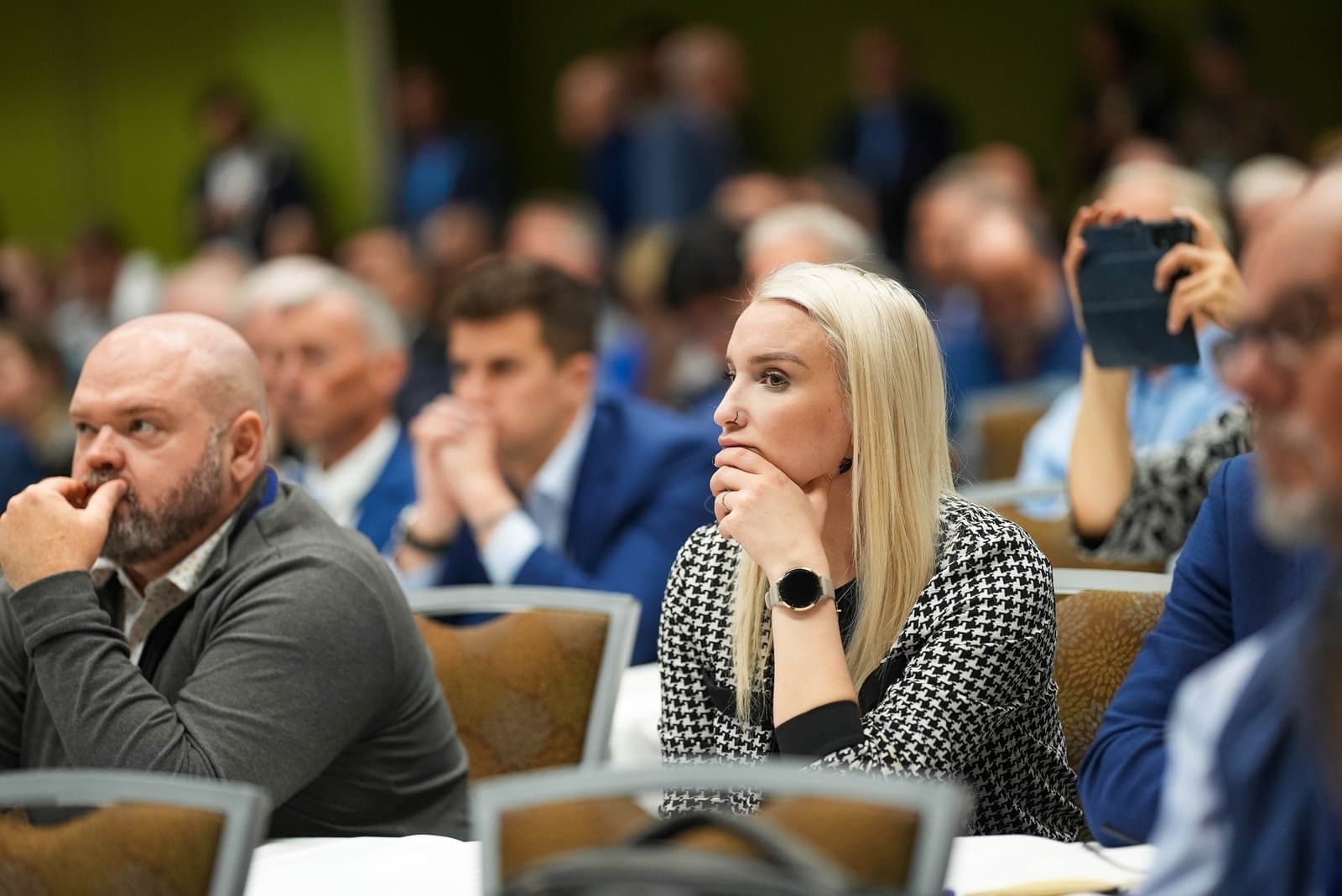 A diverse group of people, including a blonde woman with a nose ring and a bearded man, are seated and attentive at an indoor conference in Anchorage, Alaska.