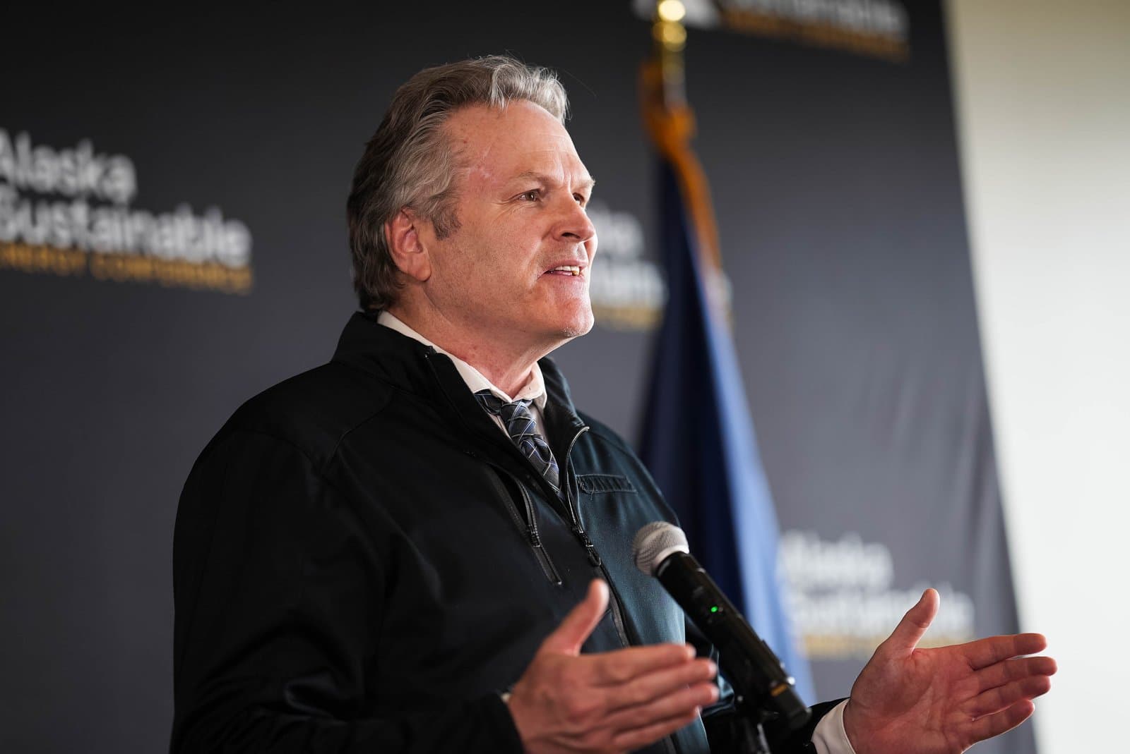 A man with gray hair, wearing a black jacket, white shirt, and patterned tie, speaks into a microphone at a podium. Behind him, a dark banner reads 'Alaska Sustainable' and a blue and gold flag is partially visible.