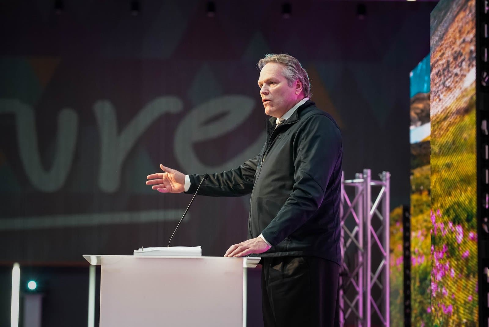 A man in a dark jacket and white shirt speaks at a podium, gesturing with his left hand. Behind him, a large screen displays part of a word, possibly 'Future', and another screen shows a vibrant natural landscape.
